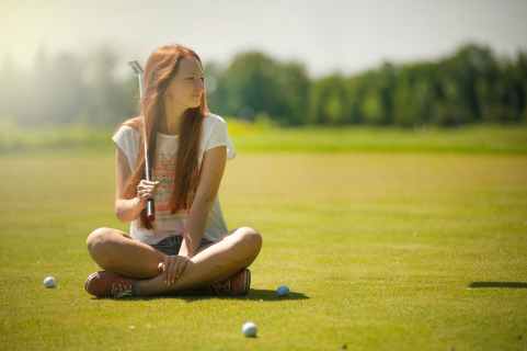 woman in white scoop neck shirt and blue shorts holding a golf club sitting on golf field